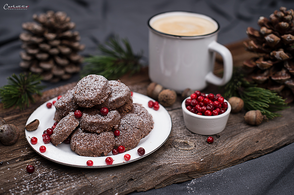 Schokoladen-Cookies mit Bröseln, Staubzucker auf weißem Teller mit Preiselbeeren auf Brett mit Kafee